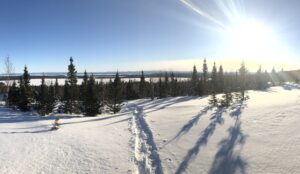Image prise du haut d'une colline enneigée. On aperçoit au loin des arbres et une étendue d'eau. Il y a des traces de pas dans la neige et le soleil brille.